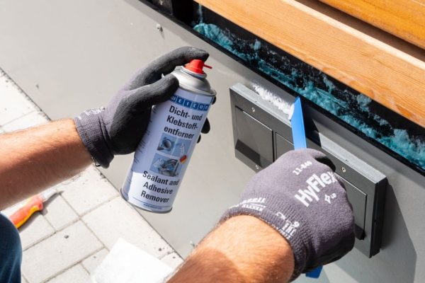 Gloved hands apply blue painter's tape to an outdoor wall panel while using a spray can labeled sealant remover nearby.