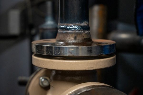 Close-up of a metal pipe joint with a circular flange and welded seam, likely part of a valve or plumbing system (industrial hardware).