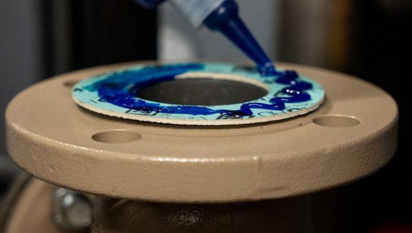 Close-up of blue adhesive being dispensed onto a circular gasket on a beige machine part.