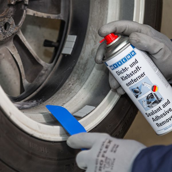 Gloved hand pressing a red nozzle spraying adhesive remover into a washing machine drum rim.