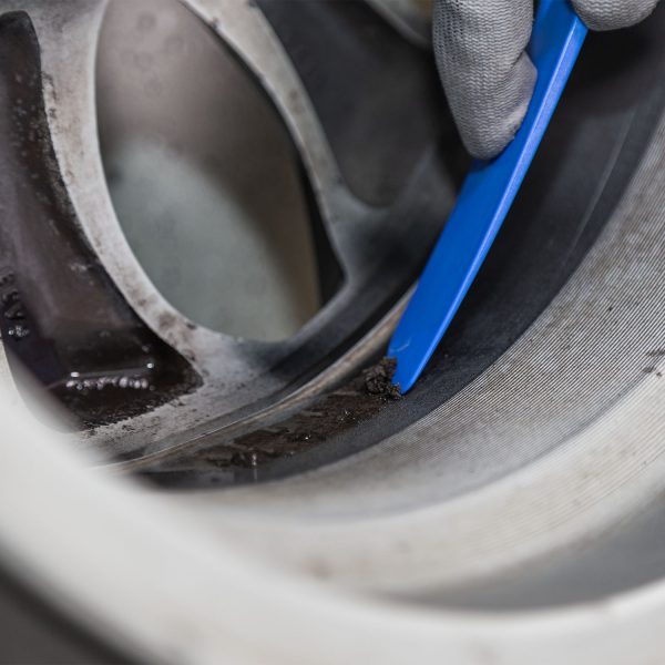 Close-up of a gloved hand using a blue plastic pry tool to lever along the edge of a circular metal component with dark grime nearby.