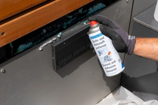 Person wearing a glove uses a spray can labeled 'Sealant and Adhesive Remover' on a gray surface near a wooden bench edge, removing sealant.