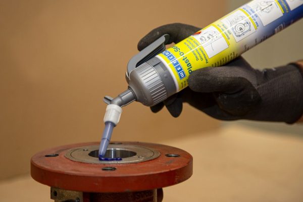 Worker wearing gloves applying blue sealant from a caulk gun into a metal flange joint on a pipe