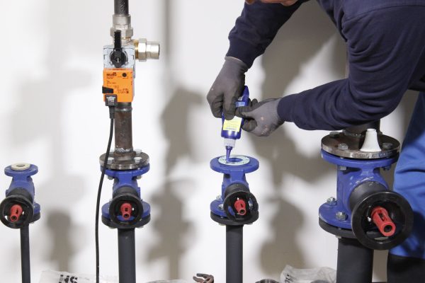 Technician in gloves applying sealant to a blue valve in a pipe system, with an orange actuator nearby.